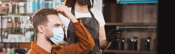 Mann in medizinischer Maske berührt seine Haare in der Nähe von Friseur in Schutzausrüstung zeigt mit dem Finger, Banner — Stockfoto