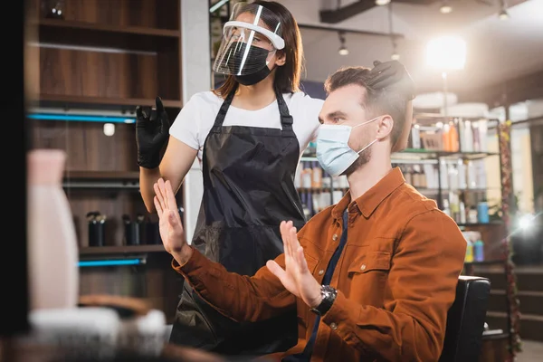 Hairdresser in protective equipment touching hair of man in medical mask showing refuse gesture — Stock Photo