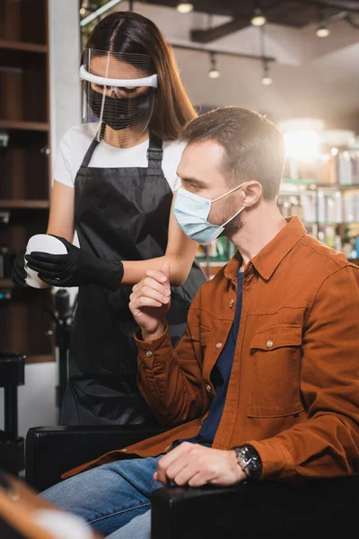 Barbeiro no rosto escudo segurando recipiente com bálsamo de cabelo perto do cliente em máscara médica — Fotografia de Stock