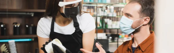 Peluquero en el protector de la cara que sostiene el envase con el bálsamo del pelo cerca del hombre en la máscara médica que señala con el dedo, bandera - foto de stock