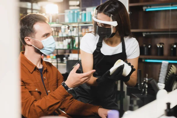 Mann mit medizinischer Maske zeigt mit der Hand in der Nähe des Friseurs in Schutzausrüstung Behälter mit Haarbalsam, verschwommener Vordergrund — Stockfoto