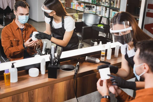 Specchio riflesso del parrucchiere in viso scudo mostrando contenitore con balsamo per capelli al cliente in primo piano sfocato — Foto stock