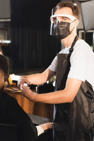 Peluquero en el escudo facial cortar el pelo del cliente mientras mira a la cámara - foto de stock