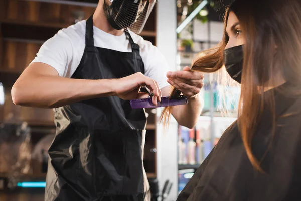 Парикмахер в фартуке и щит для лица стрижет волосы клиента в медицинской маске — стоковое фото