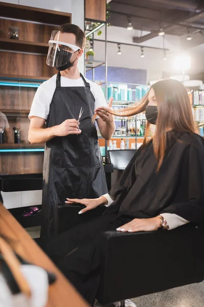 Cabeleireiro no escudo facial segurando tesoura e tocando o cabelo do cliente, foreground borrado — Fotografia de Stock