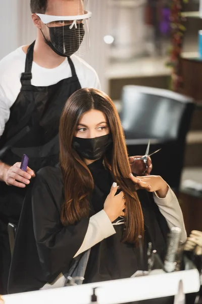 Femme en masque médical touchant ses cheveux près du coiffeur dans le bouclier facial tenant des ciseaux et peigne — Photo de stock