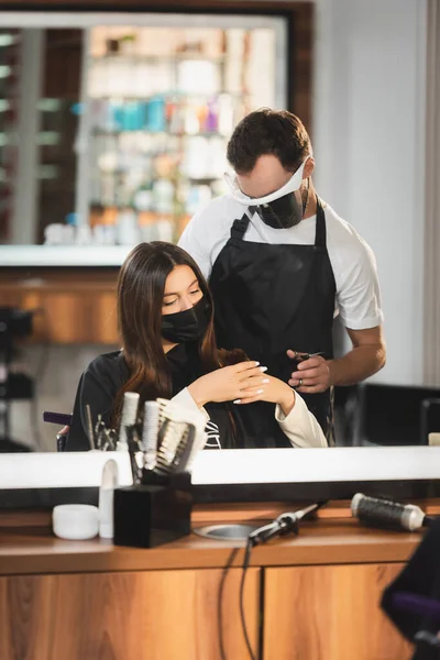 Reflejo espejo de peluquería en el escudo facial cerca de la mujer en máscara médica con herramientas de peluquería en primer plano borrosa - foto de stock
