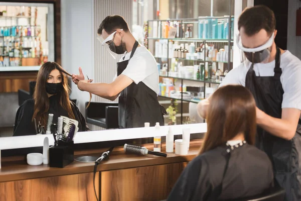 Mirror reflection of hairdresser cutting hair of client in medical mask, blurred foreground — Stock Photo