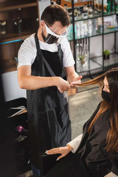 Hairstylist in face shield and apron cutting hair of woman in medical mask, blurred foreground — Stock Photo
