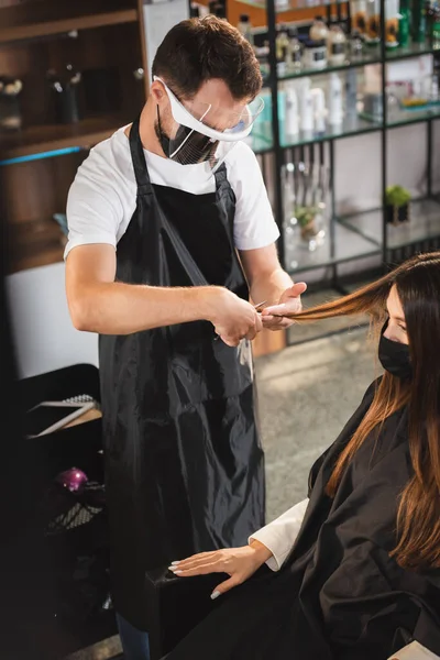 Parrucchiere in viso scudo e grembiule taglio capelli della donna in maschera medica, primo piano sfocato — Foto stock