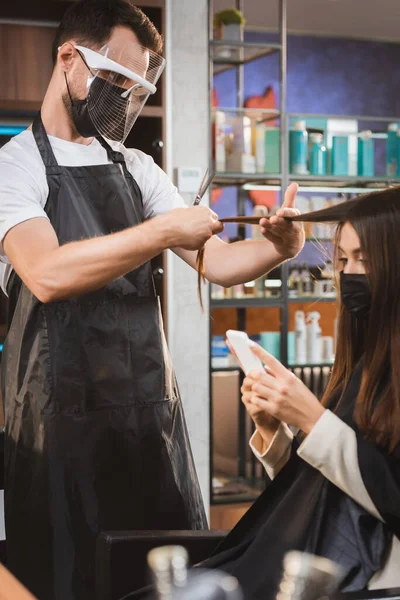 Cabeleireiro em equipamento de proteção de corte de cabelo da mulher usando smartphone, fundo borrado — Fotografia de Stock