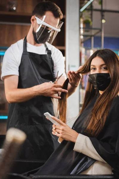 Mulher em máscara médica usando smartphone enquanto cabeleireiro no escudo facial cortando seu cabelo, foreground borrado — Fotografia de Stock