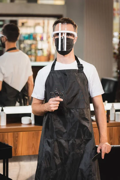 Hairdresser in face shield and apron holding scissors while looking at camera on blurred background — Stock Photo