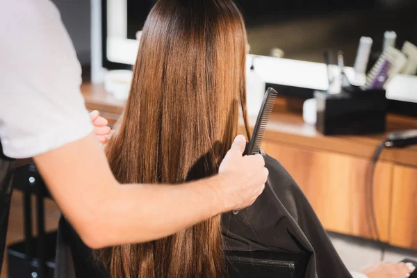 Cabeleireiro segurando pente e tocando o cabelo da mulher em primeiro plano desfocado — Fotografia de Stock