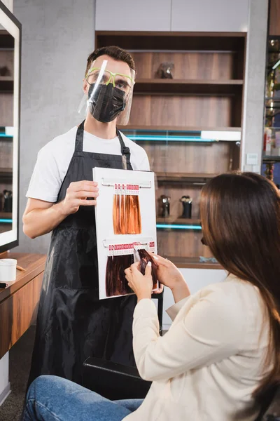 Hairdresser in face shield looking at camera and holding hair colors palette near woman touching sample — Stock Photo