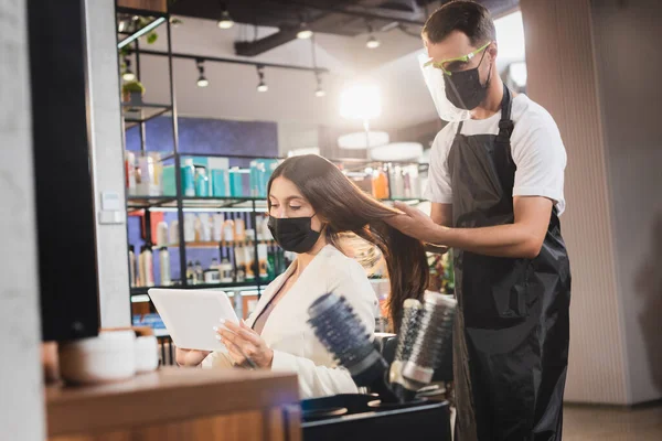 Hairdresser in face shield and apron looking at hair of woman holding digital tablet — Stock Photo
