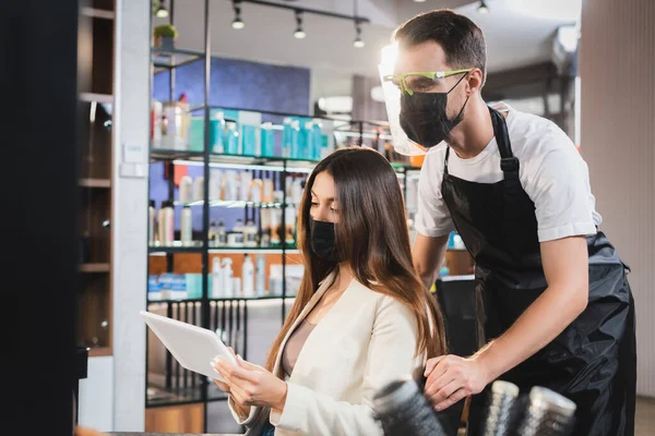 Barbiere in faccia scudo vicino alla donna in maschera medica in possesso di tablet digitale, sfondo sfocato — Foto stock