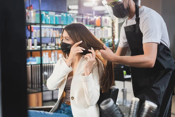 Hairstylist in apron and face shield touching hair of woman in medical mask, blurred foreground — Stock Photo