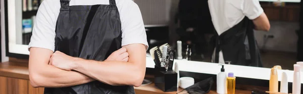 Vue recadrée du coiffeur en tablier debout avec les bras croisés sur fond flou, bannière — Photo de stock