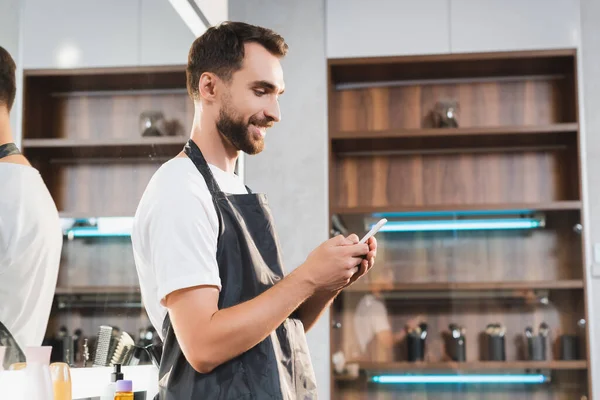 Seitenansicht eines lächelnden Friseurs, der am Arbeitsplatz mit dem Smartphone chattet — Stockfoto