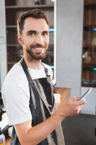 Веселый, бородатый парикмахер смотрит в камеру во время чата на смартфоне — стоковое фото