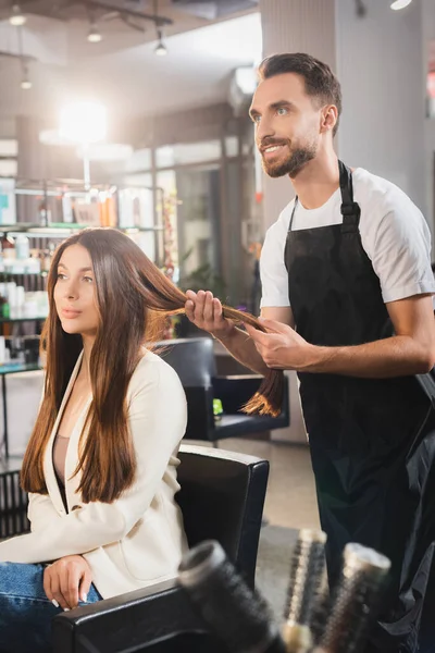 Coiffeur souriant en tablier touchant les cheveux de la jeune femme, premier plan flou — Photo de stock