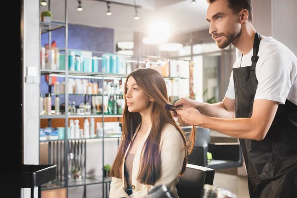Young bearded hairdresser in apron combing hair of woman — Stock Photo