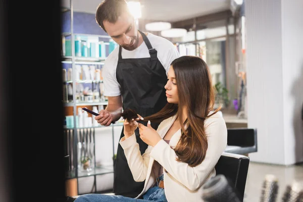 Junge Frau zeigt Friseur Haarspitzen mit Kamm, verschwommener Vordergrund — Stockfoto