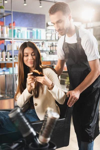 Mujer joven mostrando puntas de pelo al barbero barbudo en delantal - foto de stock