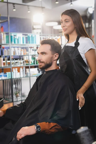 Young hairdresser in apron standing behind bearded man in barbershop — Stock Photo