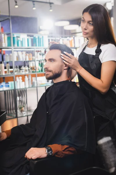 Peluquero en delantal tocando el pelo de joven barbudo en la barbería, borrosa primer plano - foto de stock