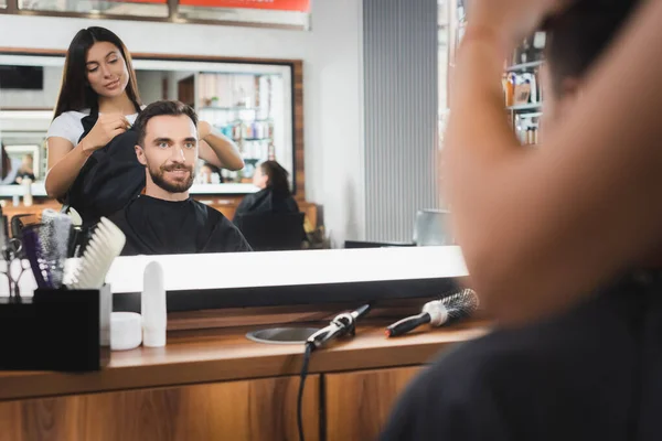 Mirror reflection of barber cutting hair of smiling man, blurred foreground — Stock Photo