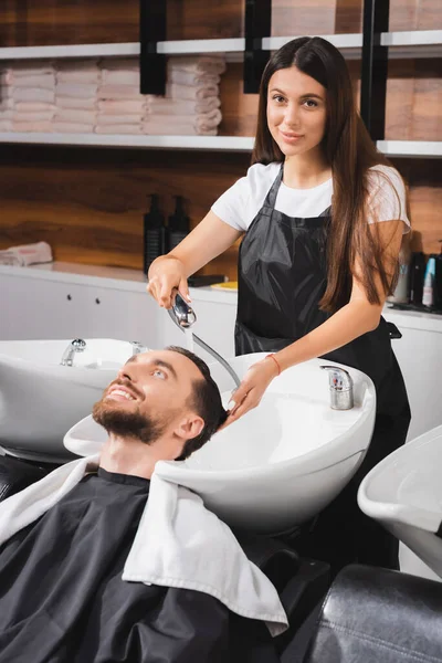 Молодой парикмахер в фартуке, смотрящий в камеру во время мытья волос счастливого клиента — стоковое фото
