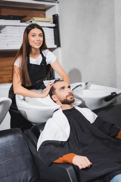 Smiling hairdresser looking at camera while washing hair of young man — Stock Photo