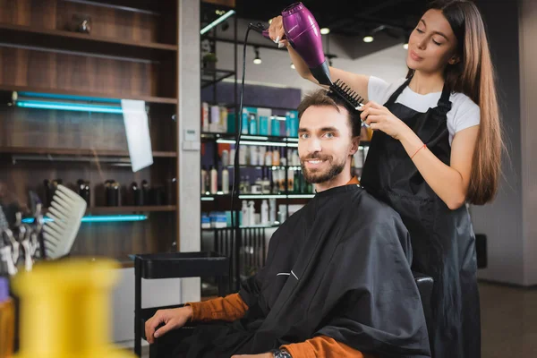 Bearded man smiling at camera while hairdresser drying his hair, blurred foreground — Stock Photo