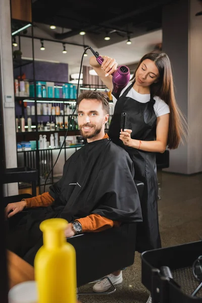 Happy bearded man looking at camera while hairdresser drying his hair, blurred background — Stock Photo