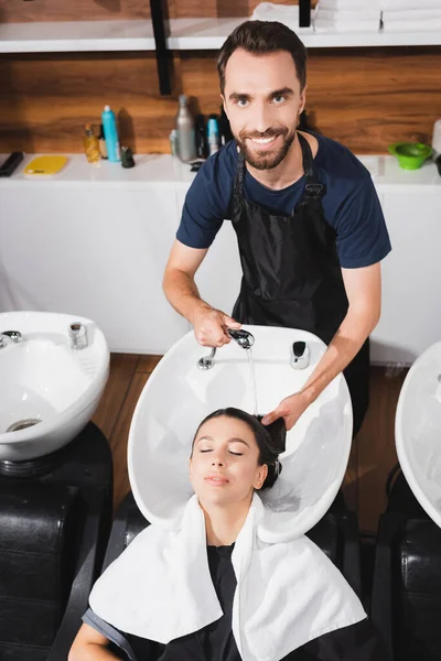 Parrucchiere sorridente, barbuto che lava capelli di giovane donna in barbiere — Foto stock