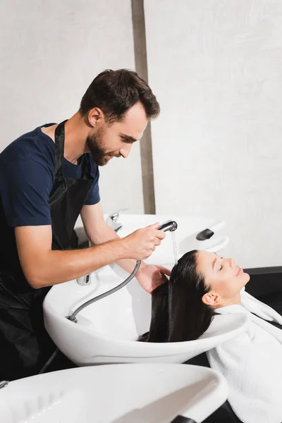 Vista laterale del barbiere barbuto che lava i capelli della giovane donna nel salone di bellezza — Foto stock