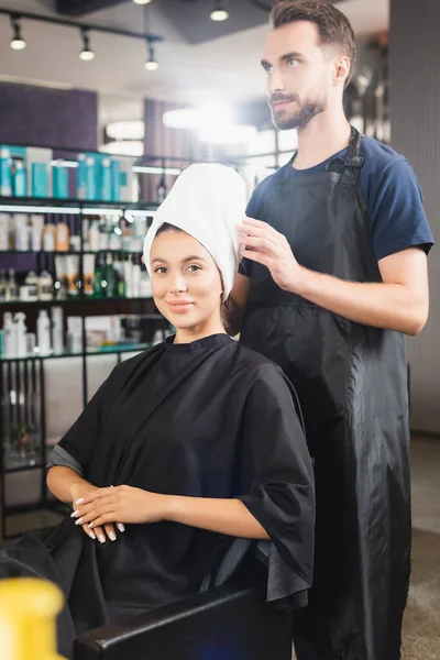 Donna sorridente con asciugamano sulla testa guardando la macchina fotografica vicino parrucchiere barbuto in grembiule — Foto stock