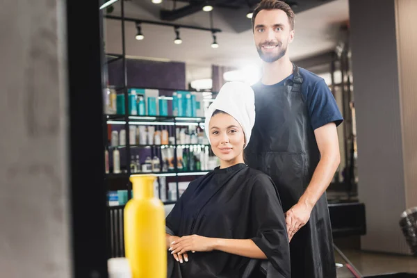 Cheerful hairdresser and smiling woman with hair wrapped in towel looking at camera, blurred foreground — Stock Photo
