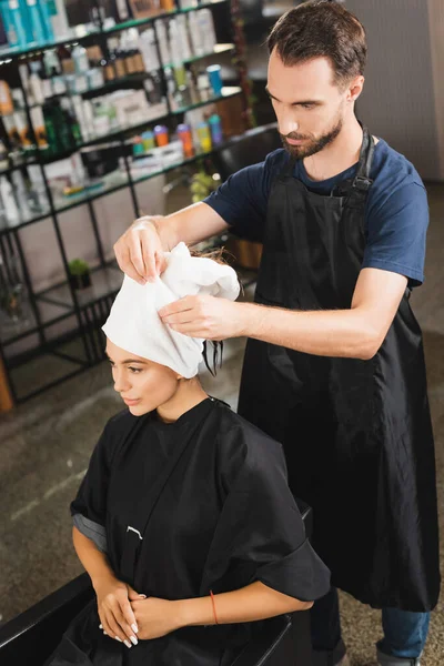 Jovem cabeleireiro barbudo no avental embrulhando o cabelo da mulher em toalha — Fotografia de Stock