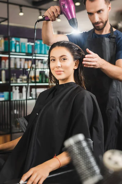 Lächelnde Frau blickt in die Kamera, während junge Friseurin ihre Haare trocknet — Stockfoto