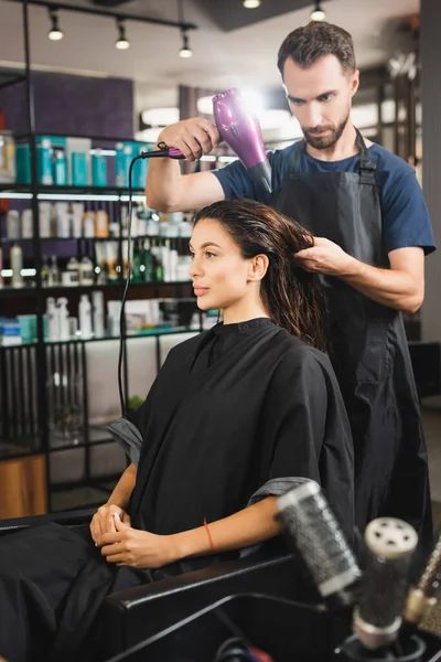 Young hairdresser drying hair of woman near combs on blurred foreground — Stock Photo