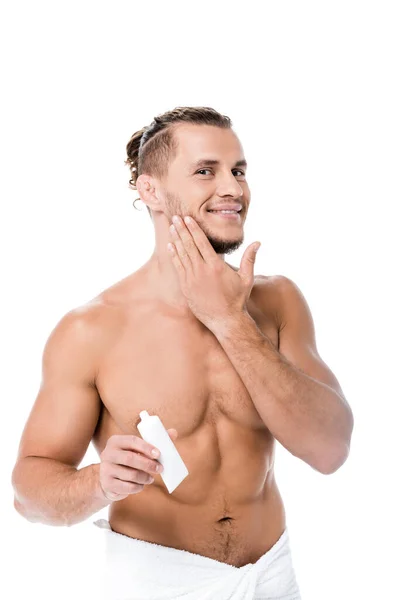 Sorrindo sexy shirtless homem em toalha aplicando creme facial isolado no branco — Fotografia de Stock