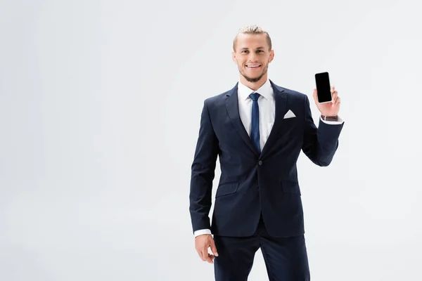 Smiling young businessman in suit presenting smartphone isolated on white — Stock Photo