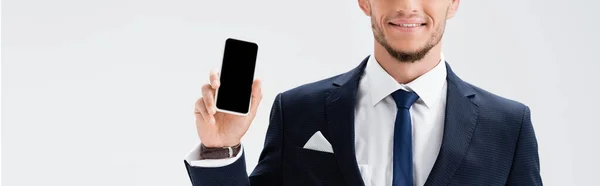 Обрезанный вид улыбающегося молодого бизнесмена в костюме, представляющего смартфон изолированный на белом, баннер — стоковое фото