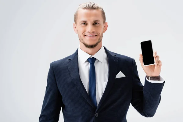 Sonriente joven hombre de negocios en traje presentando teléfono inteligente aislado en blanco - foto de stock