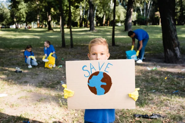 Junge hält Plakat mit Weltkugel in der Hand und speichert Aufschrift, während Familie Plastikmüll auf verschwommenem Hintergrund sammelt, Ökologiekonzept — Stockfoto