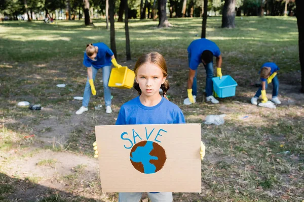 Girl holding placard with globe and save inscription, while family collecting garbage on blurred background, ecology concept — Stock Photo