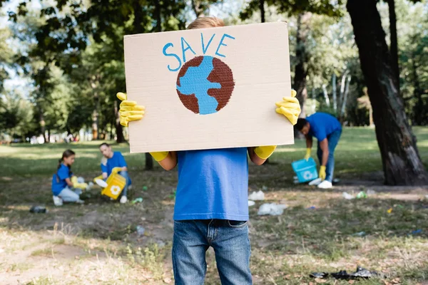 Niño sosteniendo pancarta con globo y guardar la inscripción cerca de la familia recogiendo basura en el bosque sobre fondo borroso, concepto de ecología - foto de stock